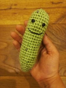 A medium green crochet pickle with a sweet smile and button eyes. It is being held up in front of a wooden background